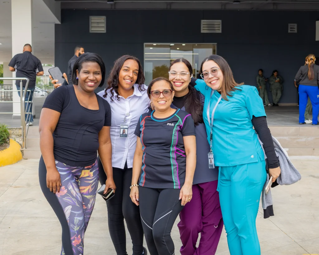 Tres colaboradores del Hospital HEMA en Santiago de los Caballeros posando sonrientes y de frente a la cámara en un set de televisión con fondo azul y el logo institucional. Al centro, un hombre con traje azul marino y corbata; a la izquierda, una mujer con chaqueta negra; a la derecha, otra mujer con chaqueta clara. Todos reflejan la energía, el profesionalismo y la integración del equipo de salud.